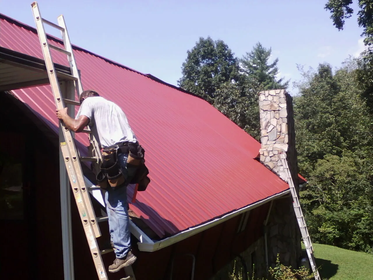 Roofer on a ladder beside a red metal roof and stone chimney — Soffit & Fascia Repair by Roofing Brothers in Sagle